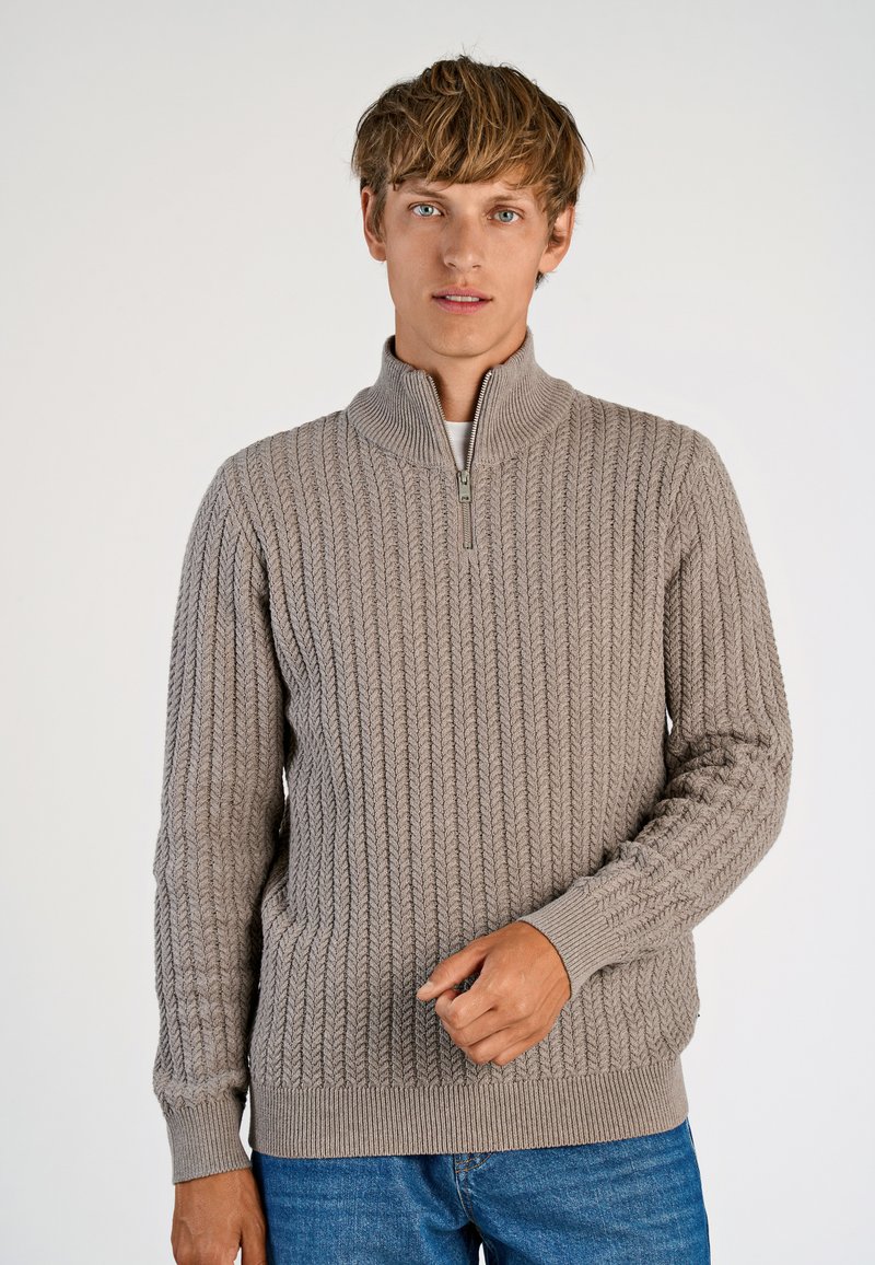 Beige Strickpullover mit hohem Kragen und halbem Reißverschluss, ausgestattet mit einem strukturierten Fischgrätmuster und gerippten Bündchen, getragen über blauen Jeans.