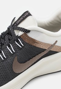 Las zapatillas Nike presentan una parte superior de malla negra, acentos texturizados en beige, suela blanca y cordones negros y blancos con puntos, además de una etiqueta de marca en la lengüeta.