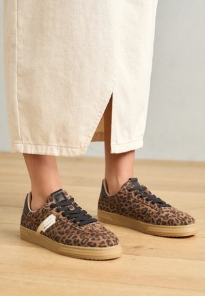 Sneakers con stampa leopardata e lacci neri indossate con gonna midi beige, in piedi su pavimento in legno.