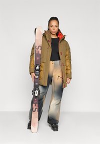 Pufi olívazöld síkabát narancssárga béléssel, színes sínadrággal párosítva. A modell síléceket tart sötét kötésekkel. Fekete sícipők láthatók.