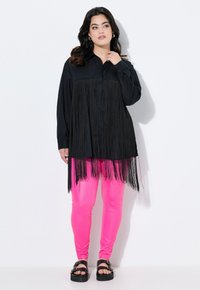 Schwarzes Hemd mit Fransenverzierung am Saum, kombiniert mit leuchtend pinken Leggings und schwarzen Sandalen mit auffälligen Schnallen.