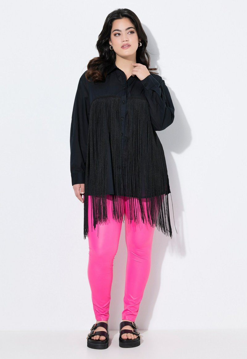 Schwarzes Hemd mit Fransenverzierung am Saum, kombiniert mit leuchtend pinken Leggings und schwarzen Sandalen mit auffälligen Schnallen.