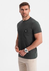 Tmavě zelené tričko s krátkým rukávem a náprsní kapsou, měkká textura, design s kulatým výstřihem a malá značka na kapse pro brandingové detaily.