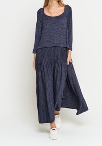 Mörkblå, långärmad topp med bred halsringning i kombination med en plisserad kjol som går ner till anklarna. Mjuk kvalitet och lös passform. Vita sneakers används.