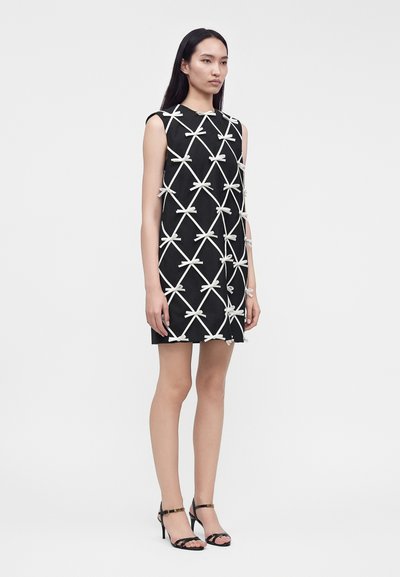 Femme portant une robe noire sans manches avec des lignes blanches croisées et des nœuds, associée à des sandales noires à talons hauts, debout sur un fond uni.