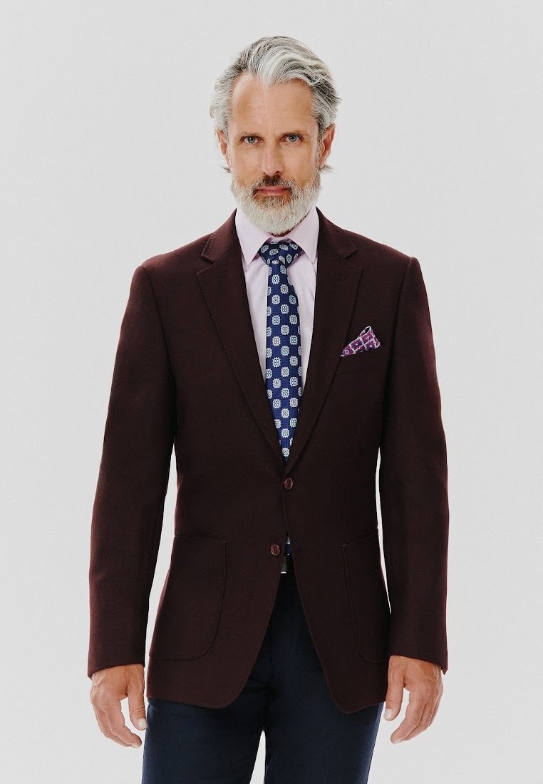 Burgundowy blazer o gładkiej fakturze, z klapami, dwoma przednimi kieszeniami i zapięciem na guziki; zestawiony z jasnoróżową koszulą i wzorzystym niebieskim krawatem.