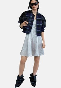 Czarna i fioletowa kurtka typu bomber z zamkiem błyskawicznym i teksturowanymi rękawami, zestawiona z jasnoszarą rozkloszowaną sukienką oraz czarnymi workowatymi botkami za kostkę.