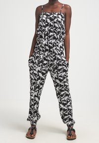 Svart jumpsuit med vitt palmbladsmönster, justerbara axelband, lös passform, fickor och elastiska ärmslut. Bärs med platta sandaler.