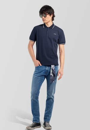 Uomo con polo nera, jeans blu con sciarpa fantasia che pende dalla tasca, sneaker nere e occhiali da sole, in piedi davanti a uno sfondo grigio.