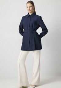 Námornícky modrá košeľa s opaskom a golierom, zapínaná na gombíky, v kombinácii s krémovými vykladanými nohavicami a svetlými topánkami.