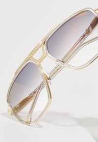 Gafas de sol de oro y plástico transparente con montura angular de gran tamaño, lentes en gradiente gris y detalle de doble puente, que cuentan con almohadillas nasales transparentes.