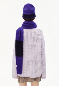 Personne portant un bonnet en tricot violet, une écharpe violette et noire assortie, un pull en laine torsadé lavande et une jupe marron, de dos.
