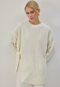 Pull oversize couleur crème avec motif en maille torsadée et poignets côtelés. Présente un col rond et des fentes latérales pour plus de confort.