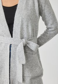 Pilka megzta kardiganas su raišteliu juosmenyje, gumbiniu rankogalių ir kišenėmis; minkšta tekstūra ir atvira priekinė išvaizda.