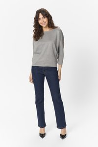 Kvinde står og smiler, iført en løs grå sweater, mørkeblå jeans og sorte spidse hæle foran en hvid baggrund.
