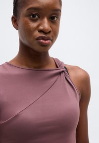 Νεαρή γυναίκα με σκουρόχρωμο δέρμα που φοράει ένα μωβ αμάνικο τοπ με ένα στριφτό λουράκι στον έναν ώμο, πάνω σε απλό φωτεινό φόντο.