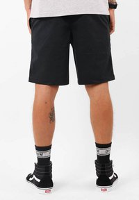 Shorts noirs au design simple, portés avec des chaussures montantes noires et des chaussettes rayées noires et blanches. Tatouage visible sur le mollet.