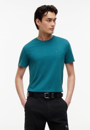 Mladý muž v tyrkysovom tričku s okrúhlym výstrihom a čiernych nohaviciach s opaskom, ruky vo vreckách, pozerá sa doprava na jednoduchom bielom pozadí.