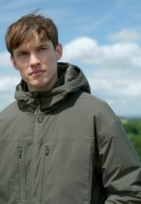 Jachetă cu glugă de culoare verde măslin, realizată dintr-un material neted, cu închidere frontală cu fermoar și două buzunare laterale. Model pozând în aer liber.