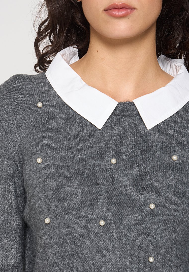 Grauer Strickpullover mit einem weißen Hemdkragen und dekorativen Perlenakzenten, die an verschiedenen Stellen auf der Vorderseite platziert sind. Weicher Stoff und lässiges Design.
