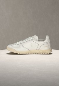 Λευκό χαμηλό δερμάτινο αθλητικό παπούτσι με μπεζ σόλα, κορδόνια και διακριτική ανάγλυφη λογότυπο στο πλάι και τη φτέρνα, σε ουδέτερο φόντο.