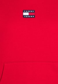 Sudadera roja con un bolsillo frontal, que presenta un parche rectangular azul oscuro en el que se lee "TOMMY JEANS" en texto blanco y rojo.