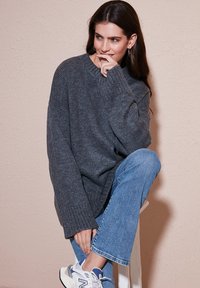Gros pull en maille gris surdimensionné avec des poignets côtelés, associé à un jean droit bleu clair et des baskets blanches, assis sur un tabouret.