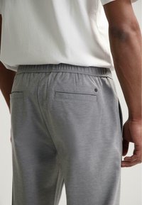 Bărbat îmbrăcat în pantaloni gri cu buzunare la spate și o cămașă albă băgată în pantaloni, prezentat din talie în jos și din lateral.