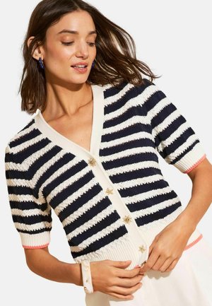 Žena v námornícky modro-bielom prúžkovanom pletenom kardigáne s hviezdičkovými gombíkmi, modrými kruhovými náušnicami a bielym náramkom, si upravuje kardigán.