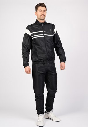 Sort trackjakke med hvide vandrette striber, lynlåslukning, elastiske manchetter, parret med matchende sorte bukser og hvide sneakers.