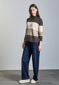 Pullover in braun, beige und cremefarbenen Streifen, mit hohem Kragen und gerippten Bündchen; kombiniert mit weiten, dunklen Jeans und hellen Sneakern.