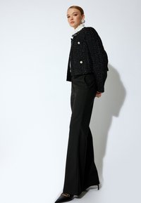 Giacca in tweed nera con tessuto testurizzato, chiusura con doppio bottone e design corto, abbinata a pantaloni neri aderenti e tacchi a punta neri.