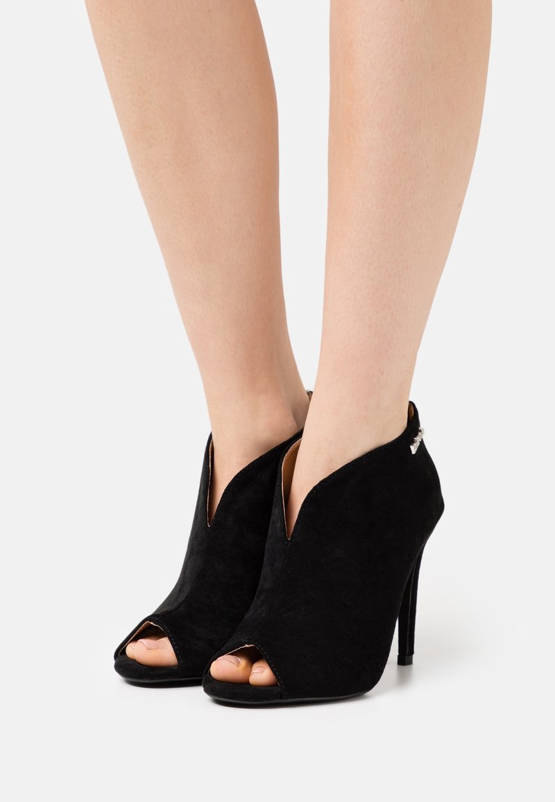 Femme portant des bottines noires en daim à talons hauts, ouvertes à l'avant avec une découpe en forme de V.