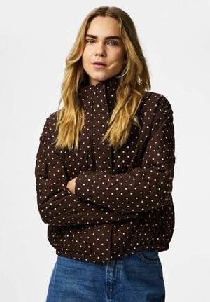 Jonge vrouw met lang blond haar, gekleed in een bruin doorgestikt jack met witte stippen en een blauwe spijkerbroek, met over elkaar geslagen armen.