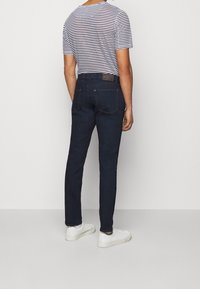 Osoba stoi tyłem do widza, ubrana w koszulę w paski oraz ciemne dżinsy, do których nosi białe trampki. Neutralne tło.