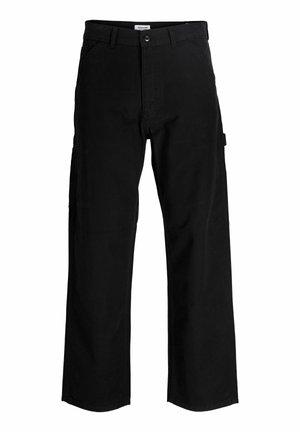 Garçon blond debout et souriant, portant une chemise crème à manches courtes avec texte et un pantalon noir ample avec des baskets blanches.