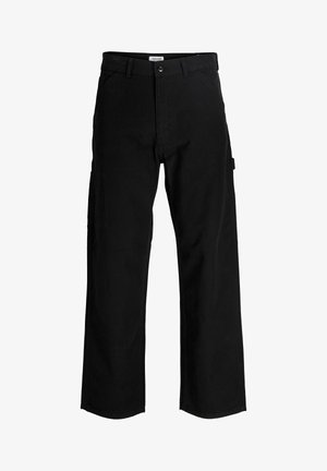 Garçon blond debout et souriant, portant une chemise crème à manches courtes avec texte et un pantalon noir ample avec des baskets blanches.