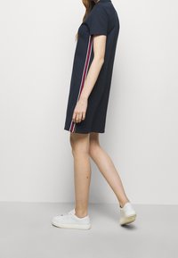 Marinblå polo klänning med korta ärmar, prydd med tre vertikala ränder i röd, vit och marinblå längs sidorna; bärs med vita sneakers.