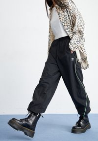 Persona caminando, vistiendo pantalones deportivos negros de Adidas con ribetes verdes, botas de plataforma negras, camiseta blanca y una chaqueta de estampado de leopardo con rayas blancas.