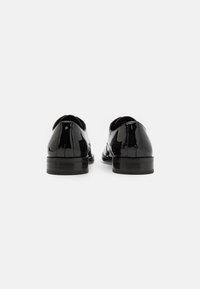 Zapatos de charol negros con puntas redondeadas, tacones bajos y una textura suave, que cuentan con costuras detalladas a lo largo de la parte trasera.
