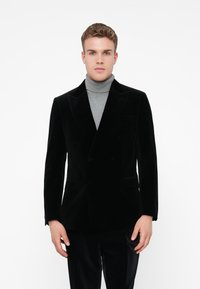 Giovane uomo con un blazer nero doppiopetto sopra un dolcevita grigio, in piedi di fronte a uno sfondo bianco uniforme.