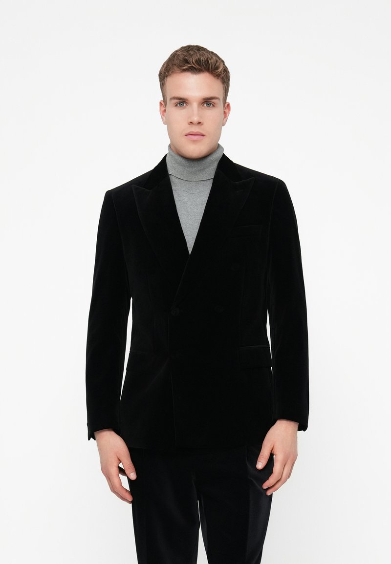 Giovane uomo con un blazer nero doppiopetto sopra un dolcevita grigio, in piedi di fronte a uno sfondo bianco uniforme.