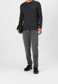 Hombre vistiendo un suéter gris oscuro con un logo amarillo, jeans grises y zapatillas negras, llevando un estuche negro de Tommy Jeans a su lado.