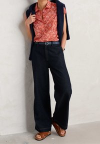 Camicetta rossa a fiori con maniche corte abbinata a jeans a gamba larga blu scuro, accentuata da una cintura blu e sandali marroni. Cardigan lavorato a maglia indossato sulle spalle.