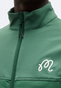 Πράσινο μπουφάν με φερμουάρ και ψηλό γιακά, με λευκό κεντημένο αφηρημένο λογότυπο στο στήθος, φορεμένο από ένα άτομο.