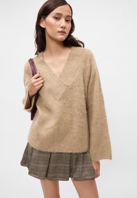 Jovem mulher vestindo um suéter bege de gola V felpudo, saias pregueadas com padrão xadrez, e segurando uma bolsa de ombro marrom-escuro, diante de um fundo branco liso.