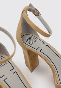 Sandalias de tacón alto de ante beiges con tacón cuadrado y tira en el tobillo, con una plantilla gris que muestra la marca "GEOX CRUZ".