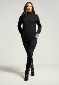 Frau steht selbstbewusst da, trägt einen schwarzen Rollkragenpullover, schwarze enge Hosen und glänzende schwarze kniehohe Stiefel vor einem schlichten hellen Hintergrund.