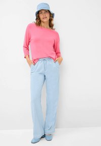 Camicia a maniche lunghe rosa abbinata a pantaloni a gamba larga a righe blu chiaro, cappello a secchiello in denim e scarpe blu. Texture morbide in tutto il look.