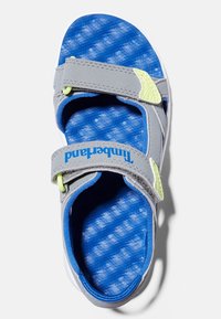 Grå sandal med blå texturerad innersula, justerbara remmar och ljusgröna accenter. Har "Timberland"-logotyp på remmen.